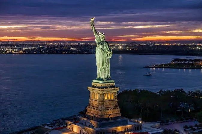 The Statue of Liberty at dusk with its pedestal floodlit and a sunset over the harbor