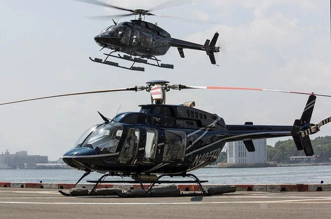 Two black tour helicopters at a waterside helipad, one lifting off